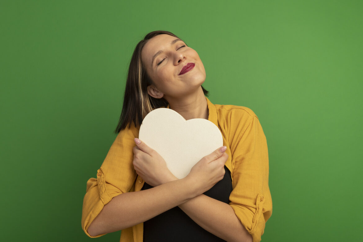 pleased pretty caucasian woman hugs heart shape isolated on green background with copy space