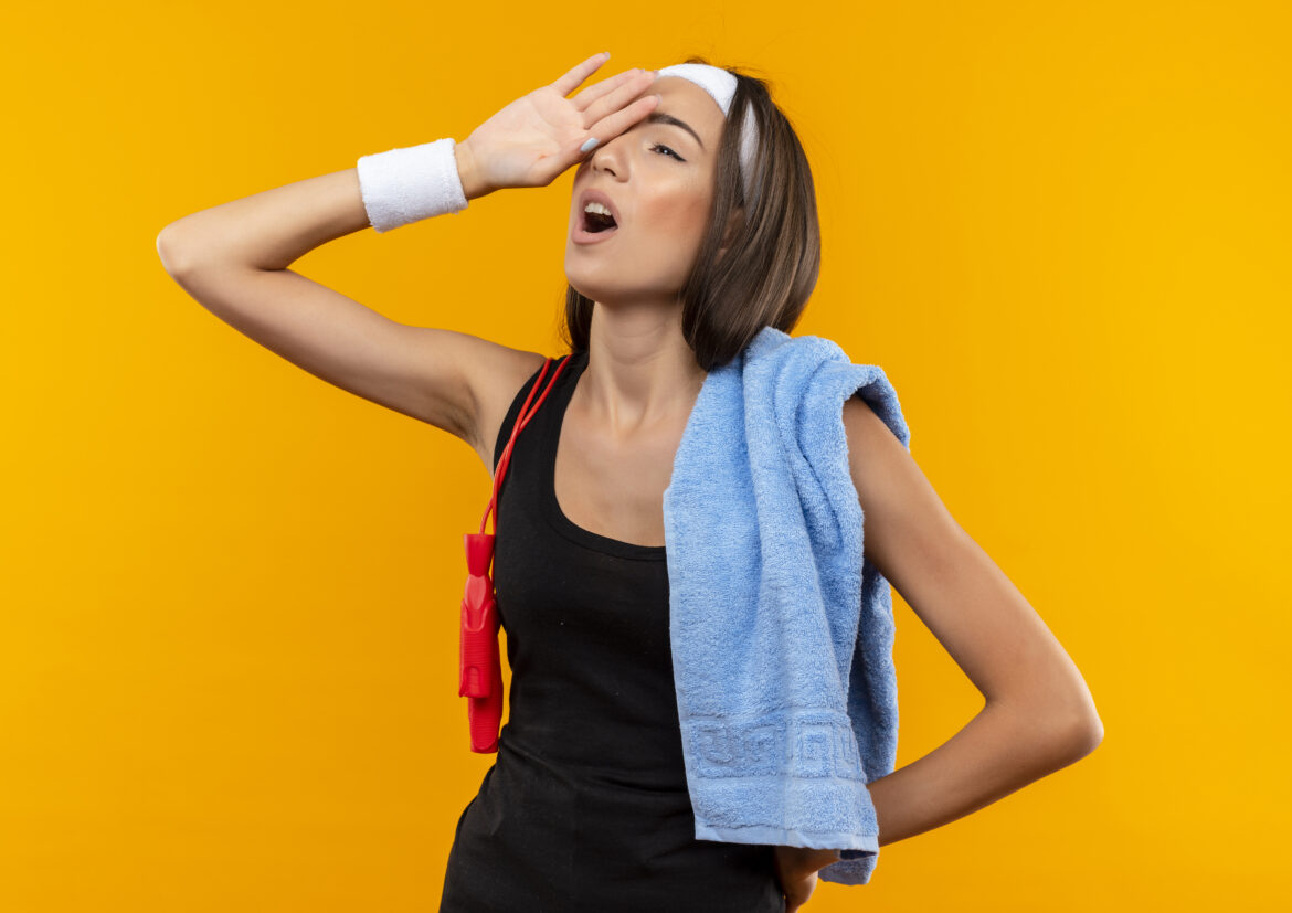 tired young pretty sporty girl wearing headband and wristband with towel and jumping rope on her shoulders putting hand on head on orange background