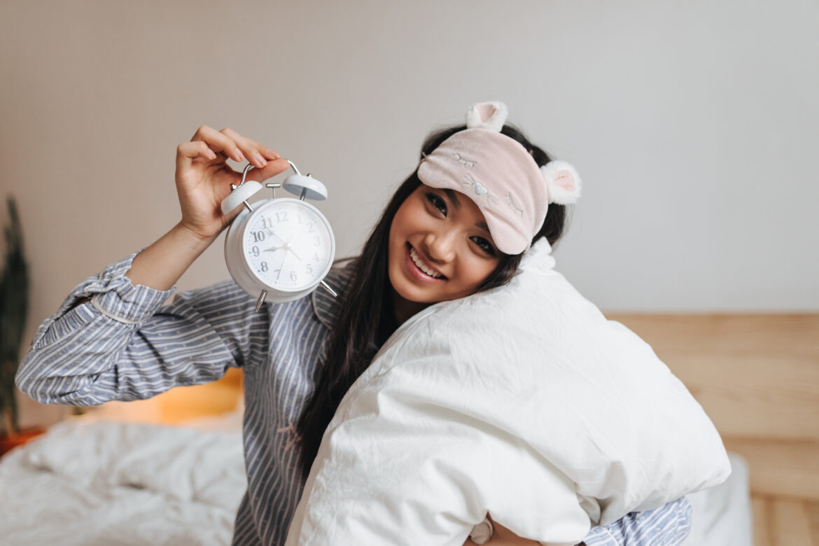 Young lady in striped pajamas keeps alarm clock. Girl in sleep mask hugging pillow