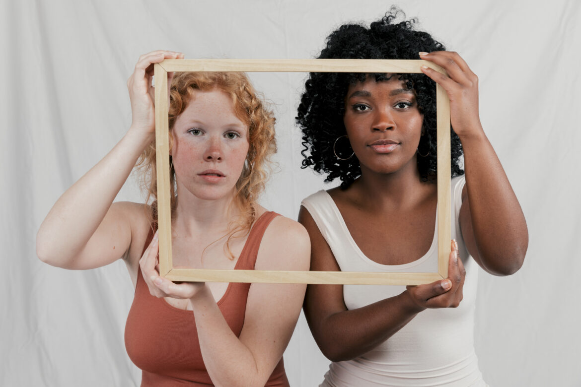 multi-ethnic-female-friend-looking-camera-through-wooden-frame-against-grey-backdrop