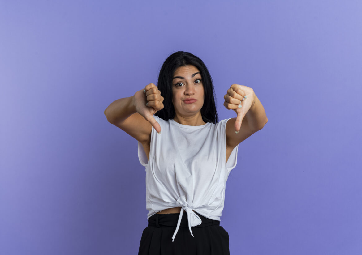 surprised young caucasian girl thumbs down with two hands isolated on purple background with copy space