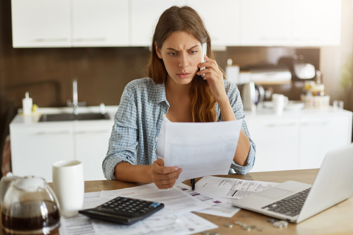 Candid shot of worried young European female dressed casually sitting at kitchen table, holding paper sheet and talking to house and utilities service representative about miscalculation in bills